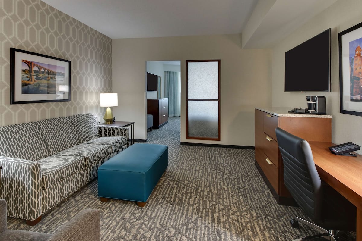 A hotel room with a patterned sofa, a blue ottoman, a desk with a computer, a lamp, and framed pictures on the walls.