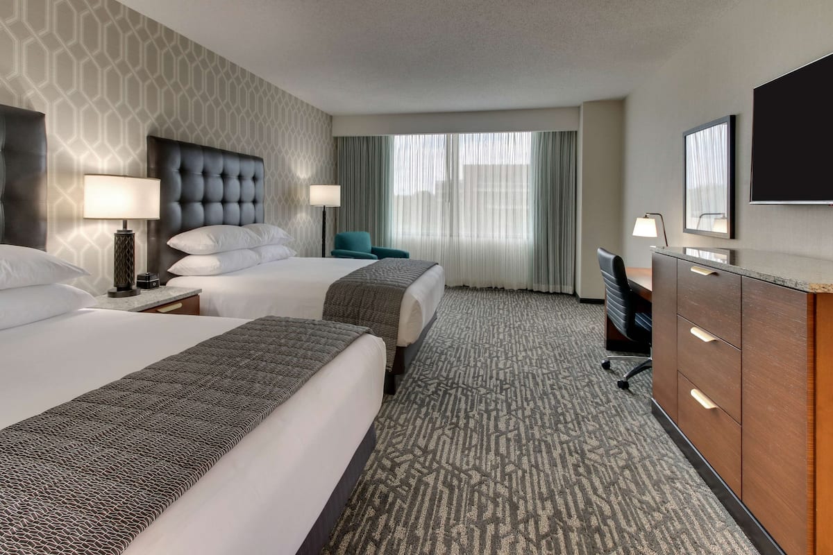A hotel room with two beds, a desk, a TV, and a window with curtains.