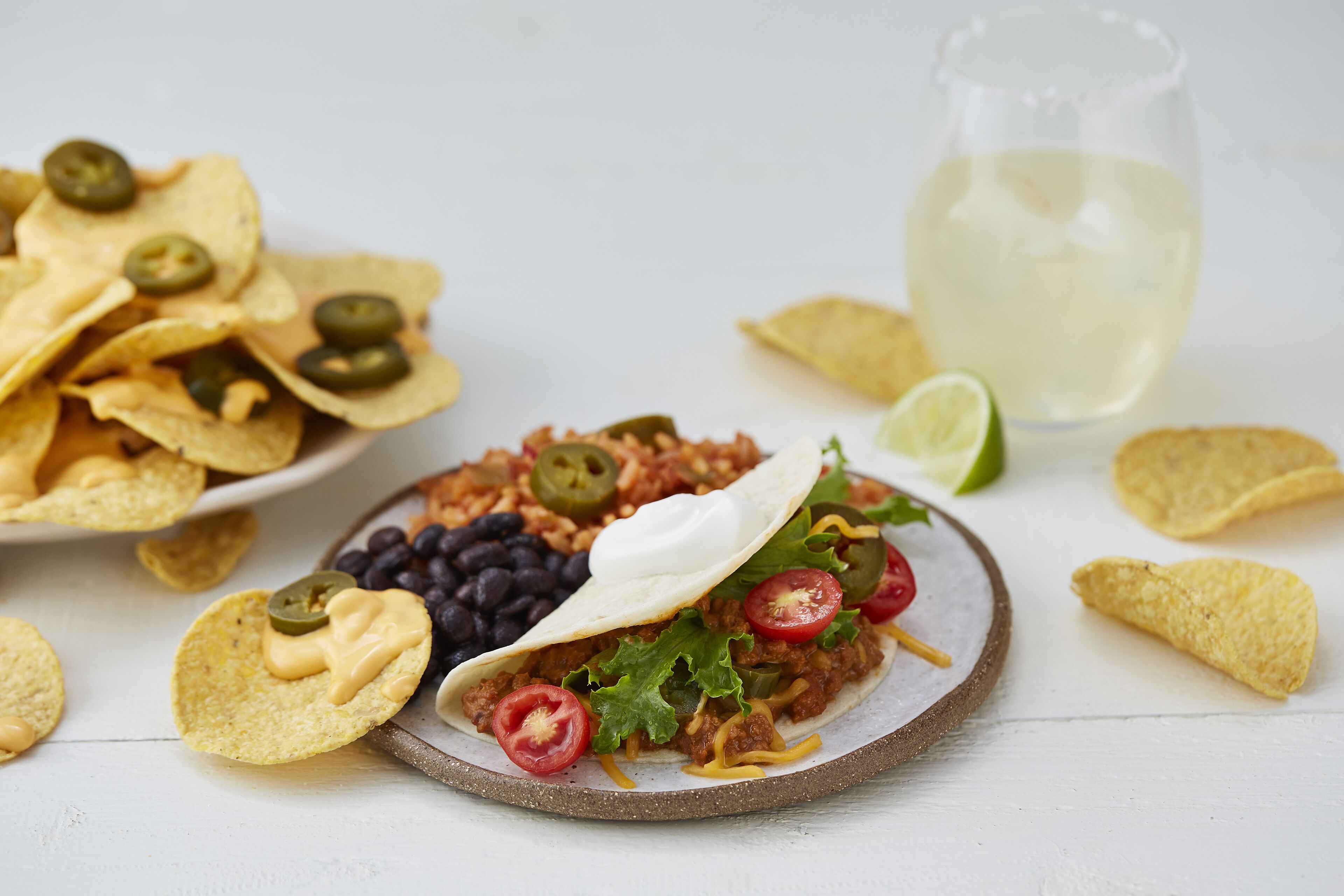 Un plato de nachos, un taco con ingredientes y un vaso de bebida blanca sobre una superficie blanca.