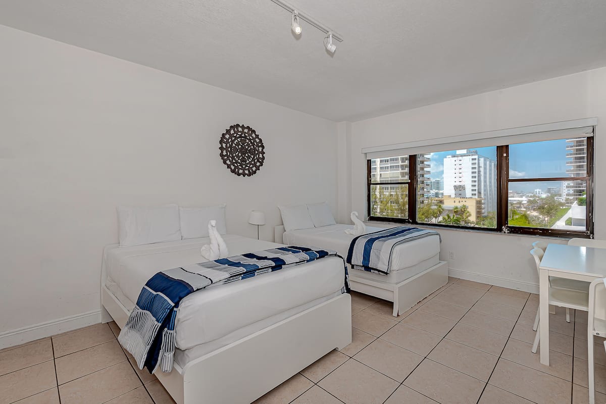 A modern bedroom with two beds, a desk, and a large window with a city view.