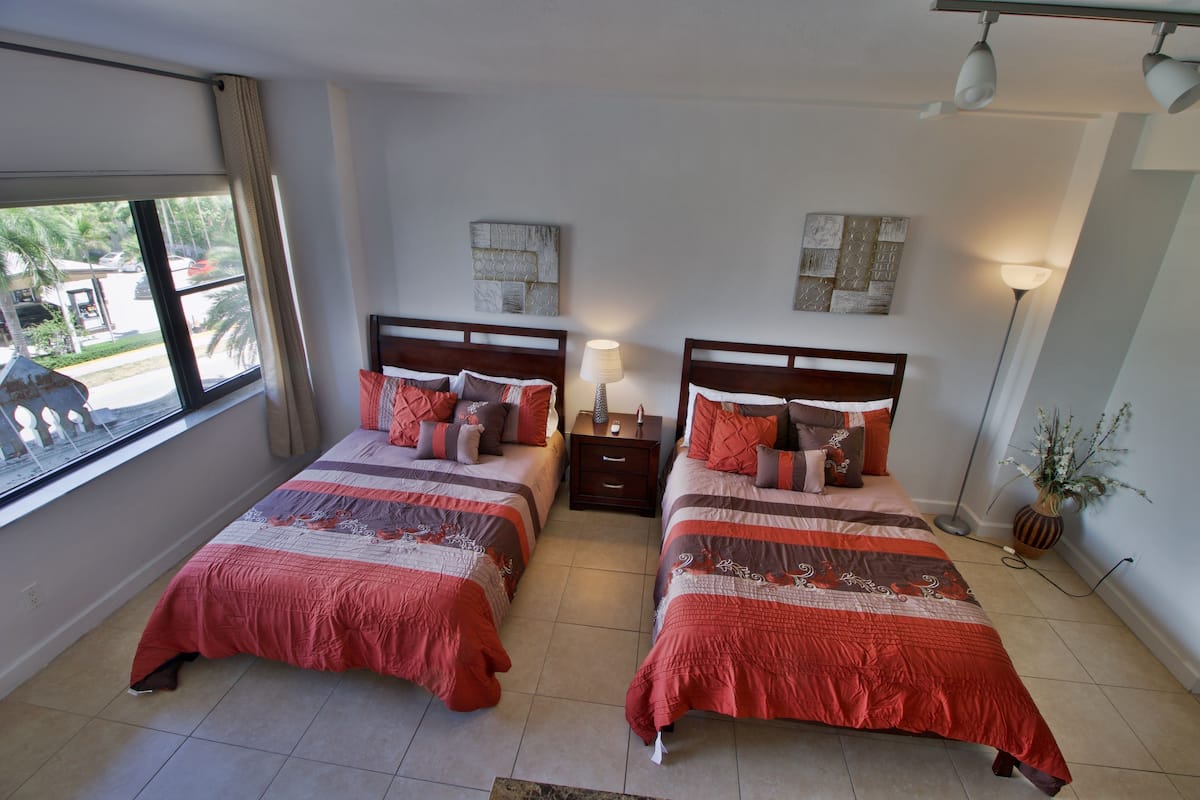 A hotel room with two beds, a window with a view of the street, a nightstand, and a lamp.