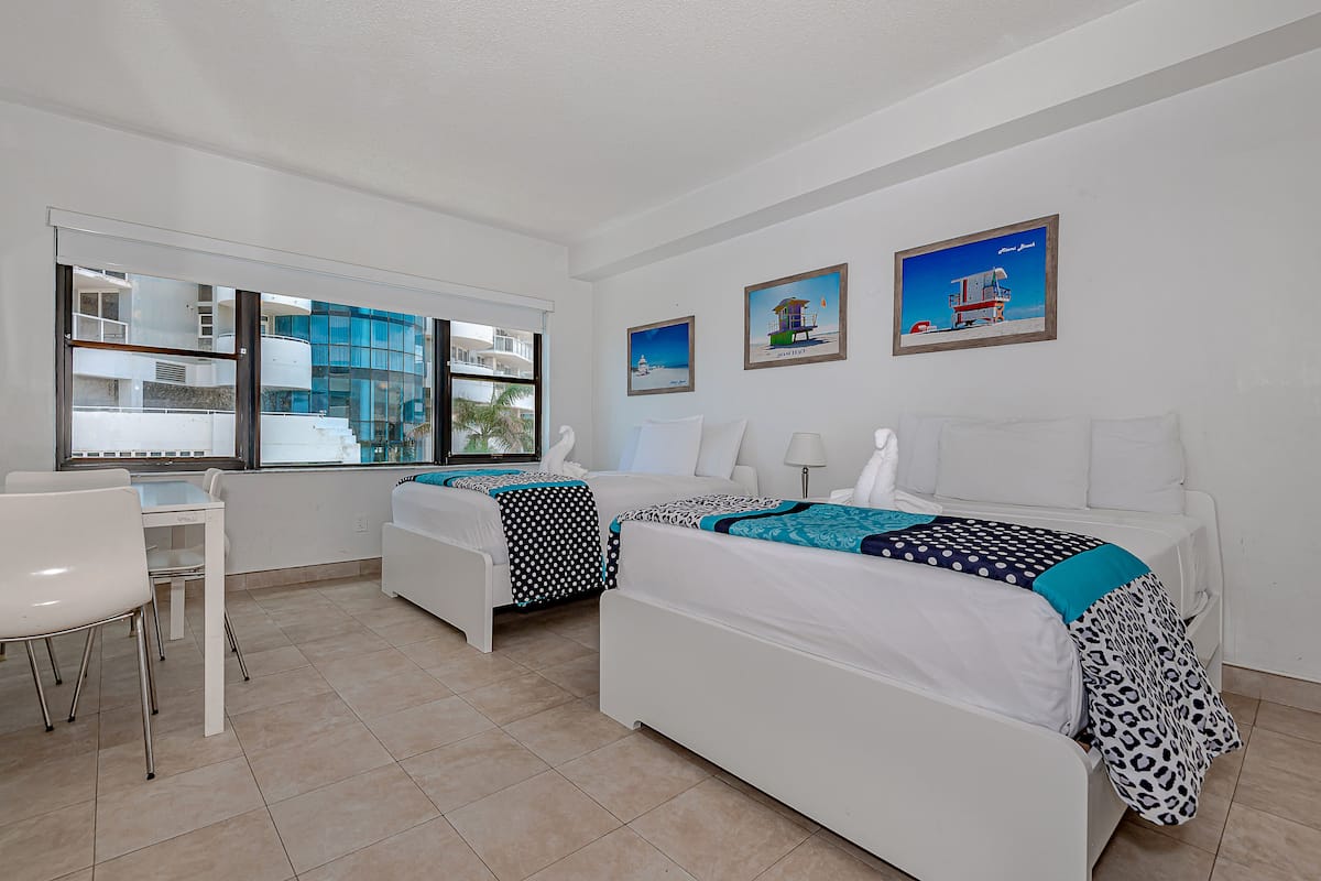 A hotel room with two beds, a dining table, and chairs. There are large windows with a view of buildings outside.