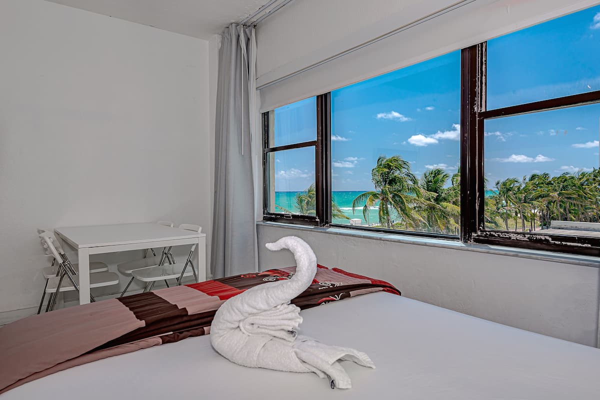 A hotel room with a bed, a desk, and a window with a view of the ocean and palm trees.