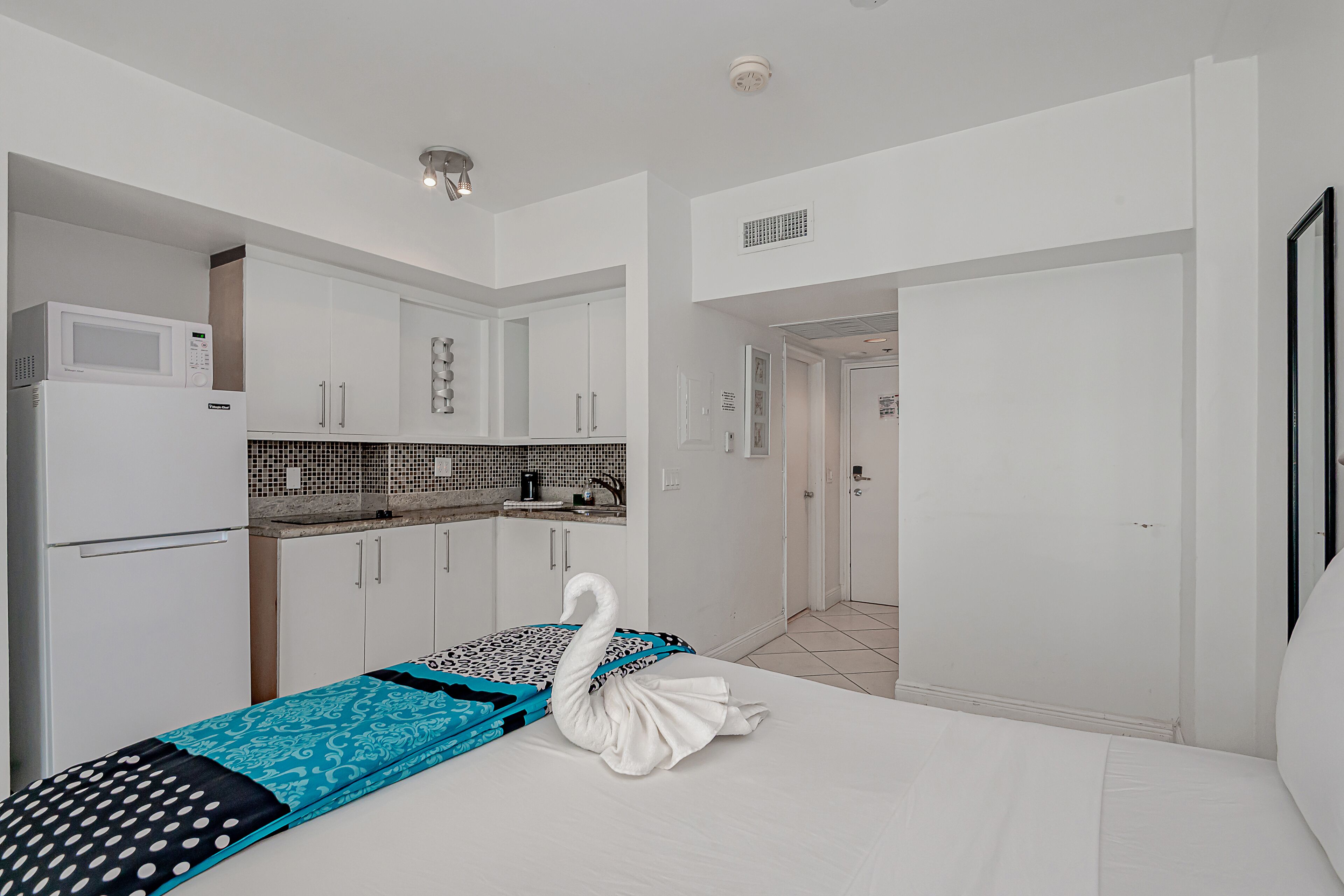 Une chambre d’hôtel compacte avec une kitchenette, un lit agrémenté d’une serviette en forme de cygne et un plan de travail carrelé dans la cuisine.