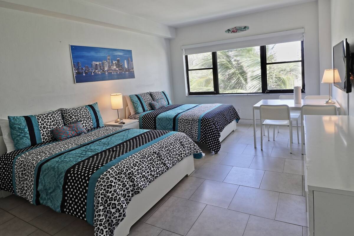 A hotel room with two beds, a desk, and a chair. There is a window with a view of palm trees and a cityscape.