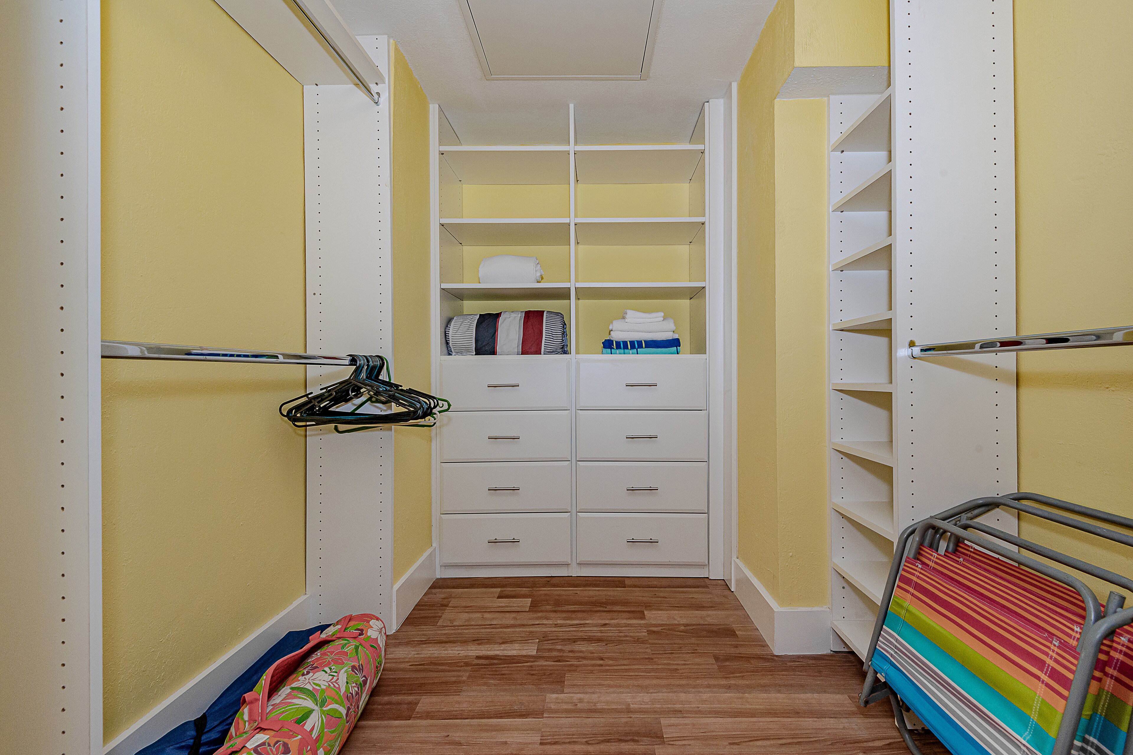 A walk-in closet with white shelves and drawers, wooden flooring, and a bench with a colorful blanket.