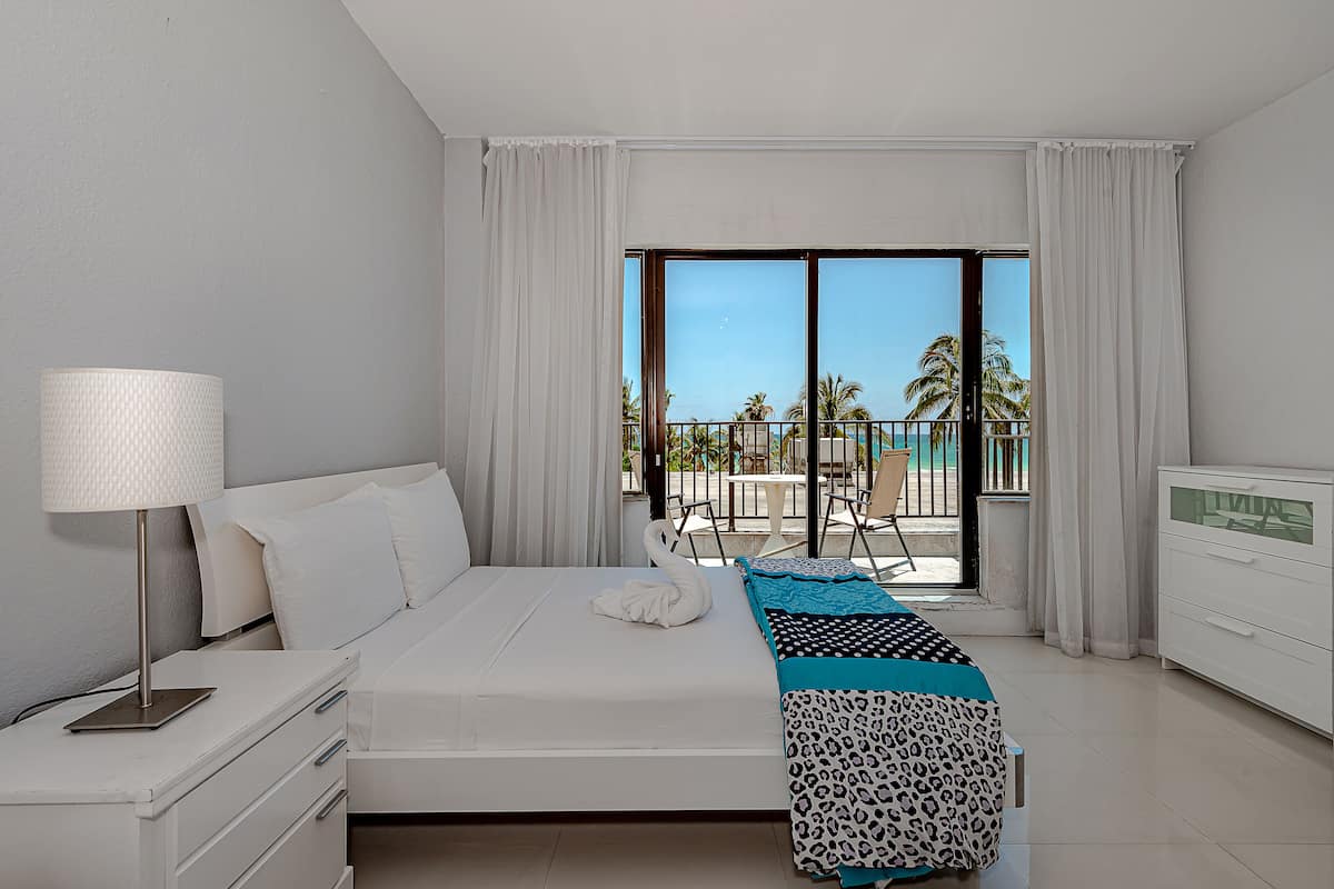 A modern hotel room with a large bed, a bedside table, a lamp, a view of the balcony, and a white dresser.