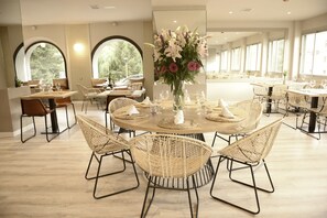 Een eetgedeelte met ronde tafels, rieten stoelen en een bloemarrangement in het midden.
