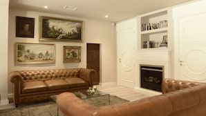 A living room with a brown leather sofa, a glass coffee table, and a fireplace. There are framed paintings on the wall and a bookshelf with books and decorative items.