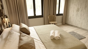 A neatly made bed with beige bedding and pillows, a rolled towel on the bed, a wicker chair, and a wardrobe with a light wood finish.