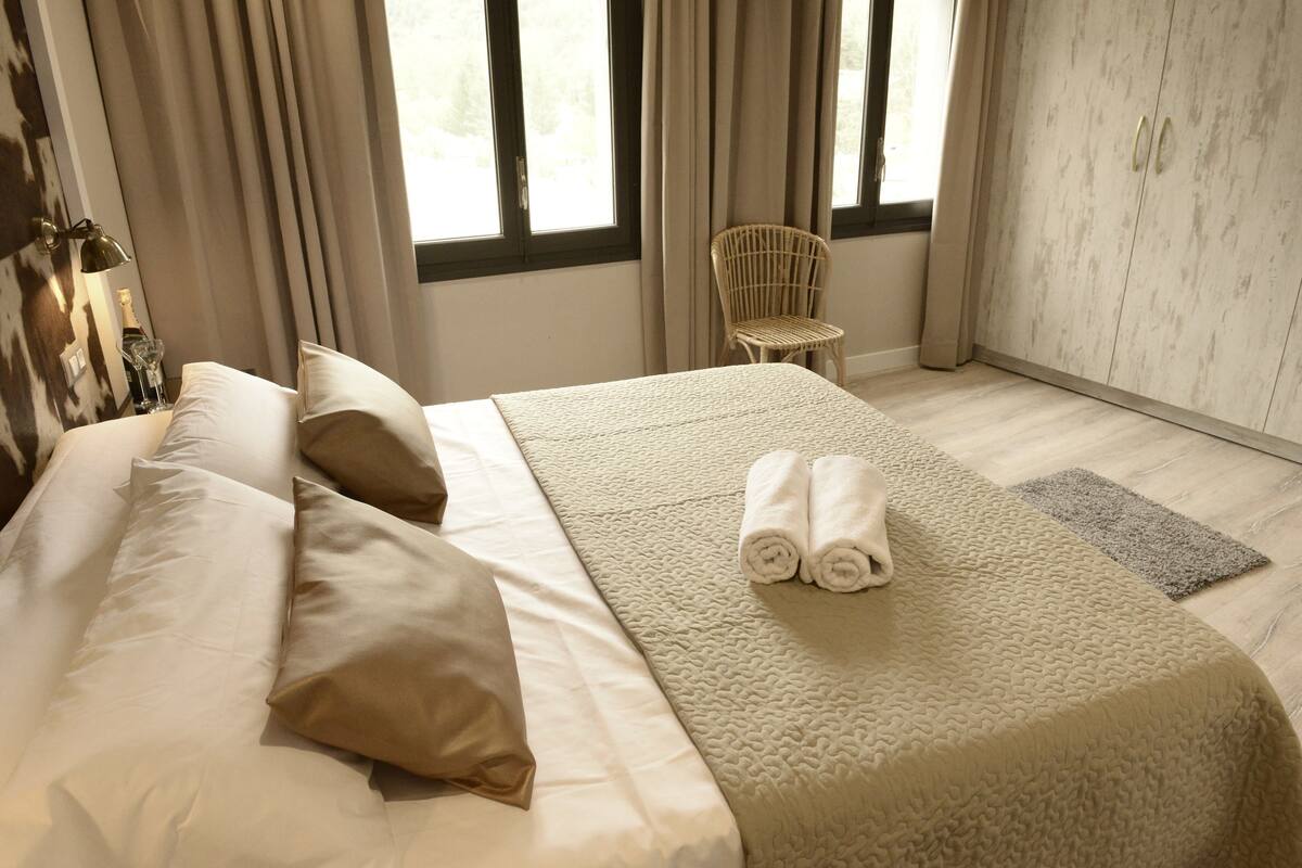 Una cama bien hecha con ropa de cama y almohadas color beige, una toalla enrollada sobre la cama, una silla de mimbre y un armario con acabado en madera clara.