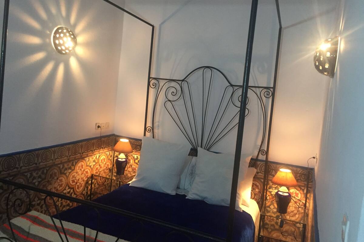 A bed with a metal headboard, two bedside lamps, patterned wallpaper, and a power outlet on the wall.