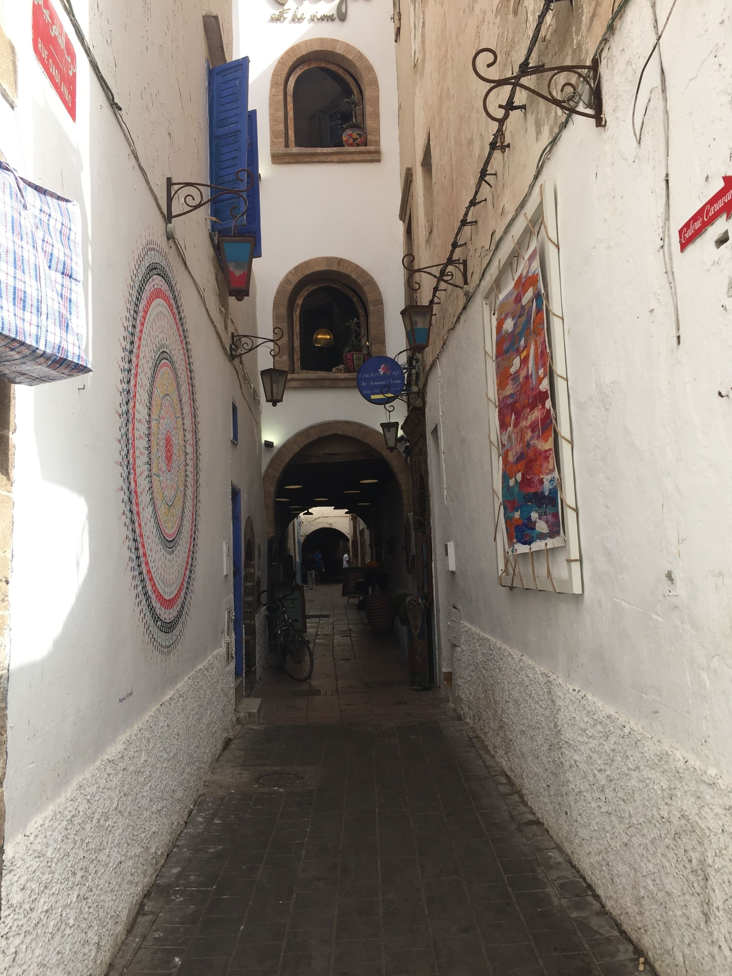 A narrow alleyway with a tiled floor, arched doorways, and a mural on the left wall.