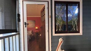 A door leading to a room with a bed and a lamp, a window with a bamboo design, and a pelican standing outside. - Pelicans Rest Shoalwater Bay
Pelican Retreat (Shoalwater)