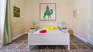 A bedroom with a white bed, two bedside tables with lamps, patterned floor tiles, and a green wall with artwork. - BLU HOUSE  (Sliema)