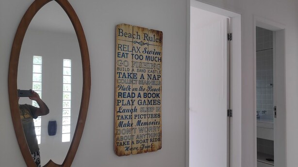 Un baño con espejo de marco de madera, un letrero en la pared con frases alusivas a la playa, y una puerta entreabierta que da a otra habitación.