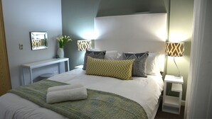 Ein ordentlich bezogenes Bett mit einer grün-weißen Decke, einem weißen Handtuch und gemusterten Kissen. Ein Nachttisch mit Lampe und Blumenvase ist vorhanden. Eine Holzgarderobe ist links im Bild zu sehen.