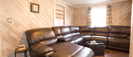 A brown leather sectional sofa with a tray table and a small side table, a window with plaid curtains, and a wooden slatted ceiling.