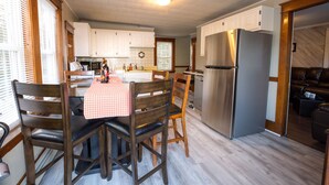 A modern kitchen with white cabinets, a stainless steel refrigerator, and a dining area with wooden chairs and a checkered tablecloth. - Sunnyside home near Sunday River, Black Mountain, Lakes, Rivers and Hikes (Rumford)