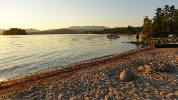 A beach with a boat on the water, a rocky shoreline, and trees in the background. - Sunnyside home near Sunday River, Black Mountain, Lakes, Rivers and Hikes (Rumford)