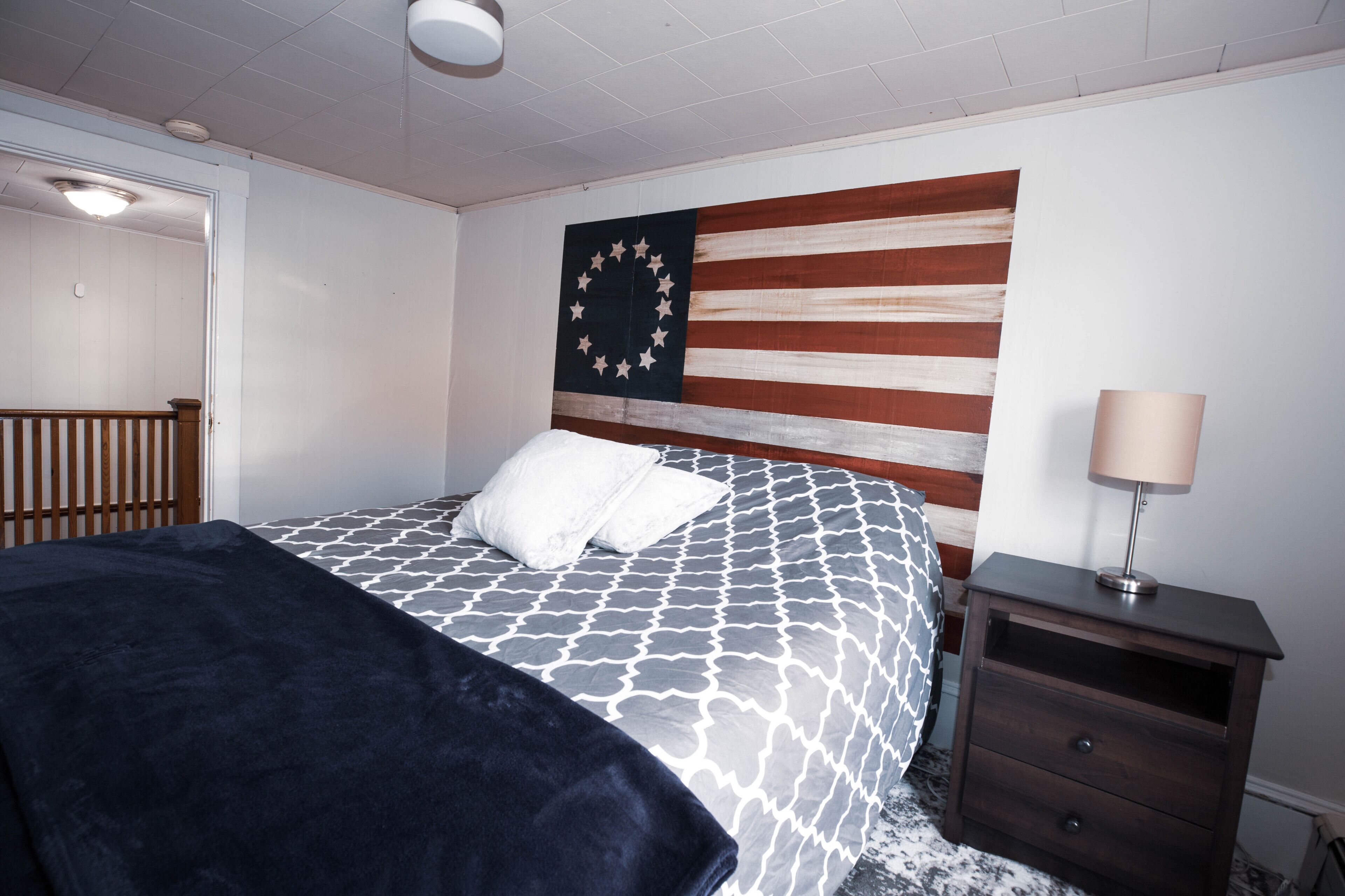 Une chambre à coucher avec un lit, une table de chevet, une lampe et un mur orné d’un grand drapeau américain.