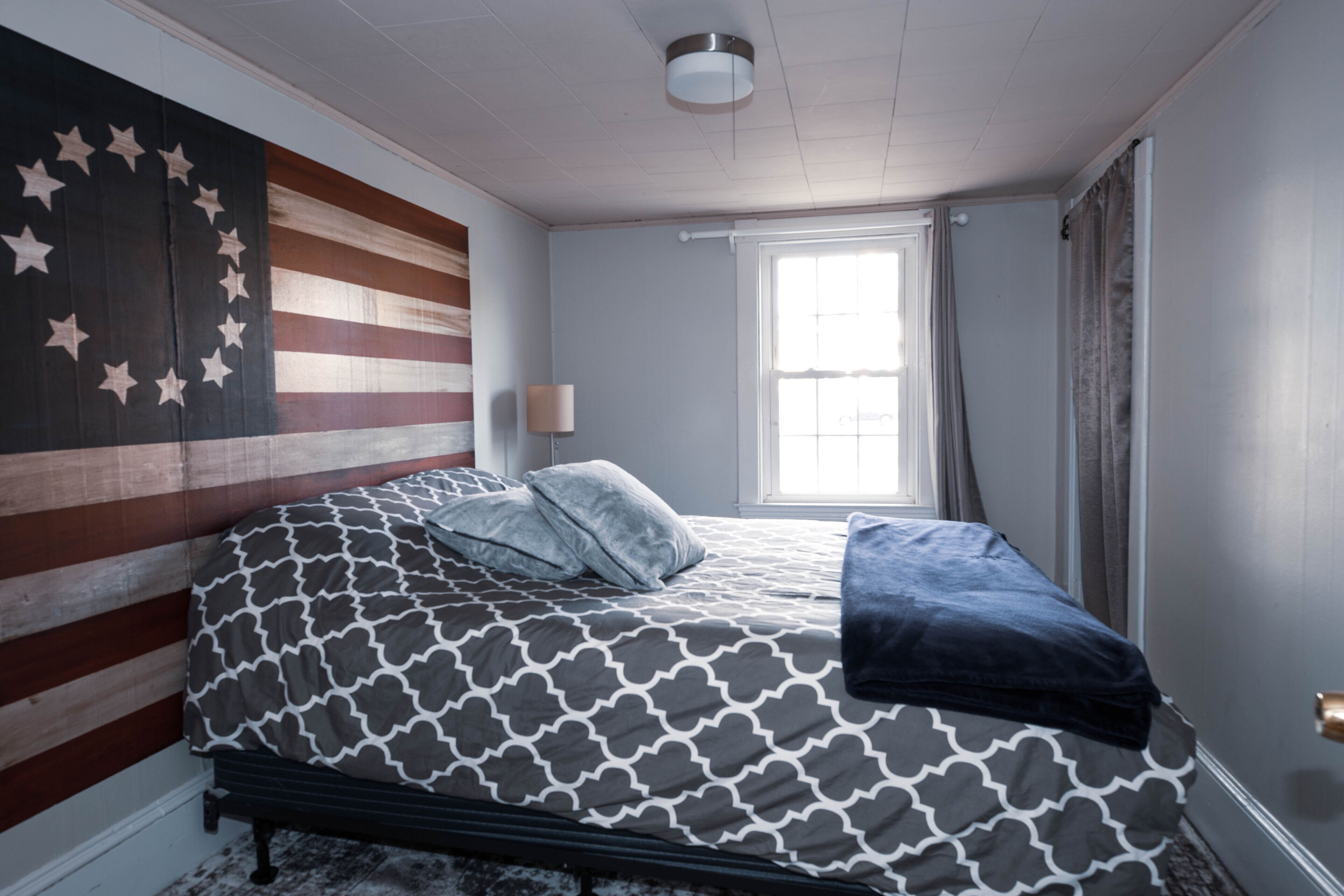 A bedroom with a bed, a large American flag on the wall, a window with curtains, and a nightstand with a lamp.
