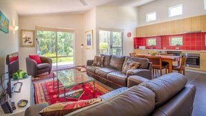 A spacious living room with a leather sofa, a glass coffee table, and a TV. There is a kitchen with red cabinets and a dining area with a table and chairs. - 2bdr villa in Dunsborough (Dunsborough)