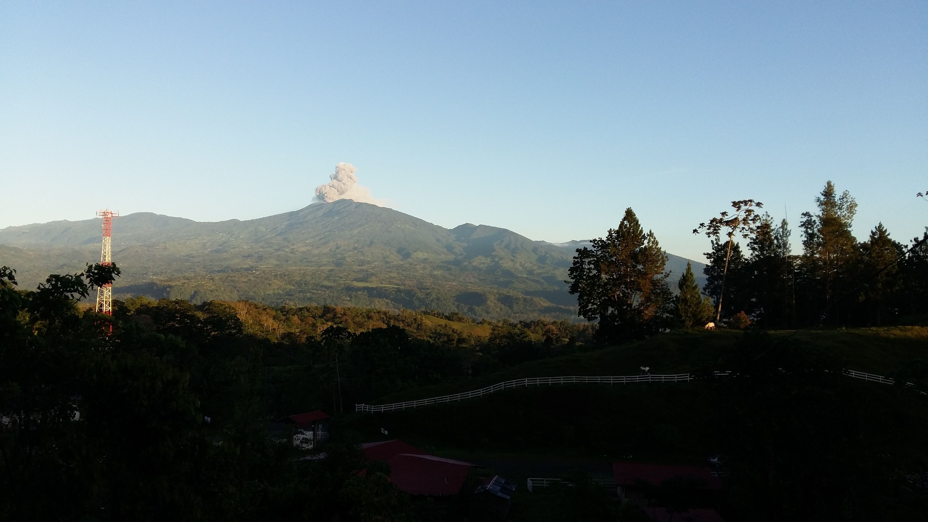 A mountain with a smoke plume, surrounded by trees and a few buildings.