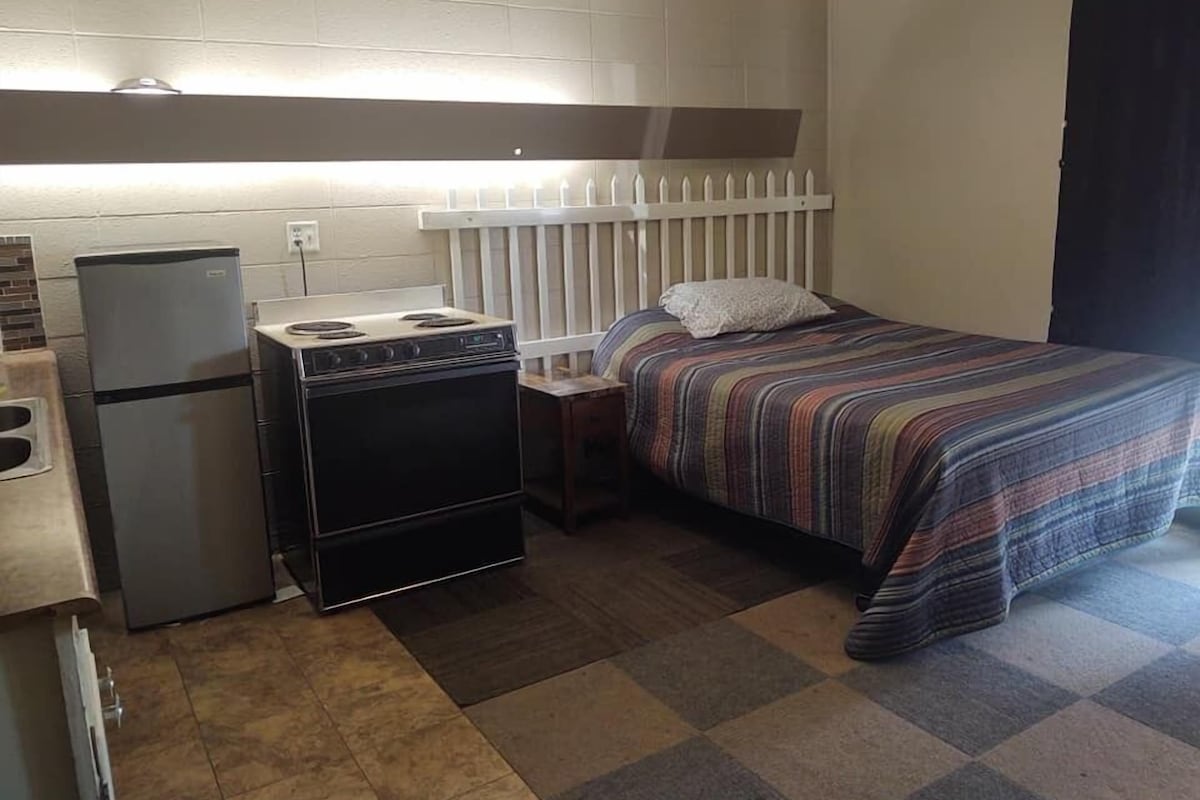 Ein kleines Zimmer mit Küchenzeile, inklusive Kühlschrank, Herd und Spüle. Ein Bett mit gestreifter Decke und Kissen vorhanden.