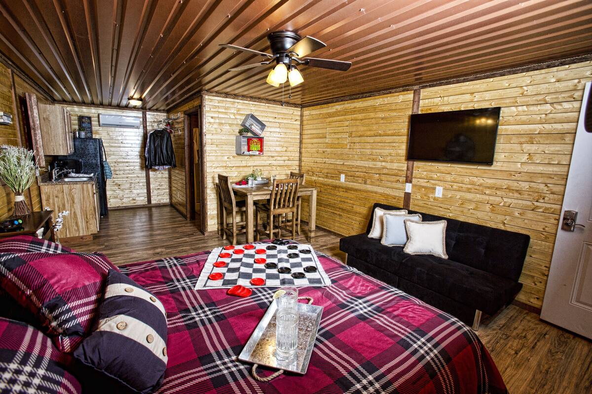 A cozy room with a checkered bedspread, a wooden ceiling, a television mounted on the wall, and a small dining area with a table and chairs.
