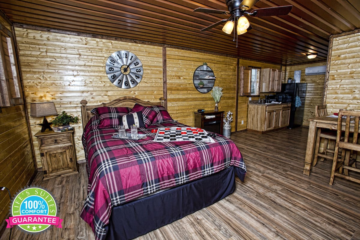 A cozy log cabin room with a large bed, a wooden ceiling fan, a kitchen area, and a clock on the wall.