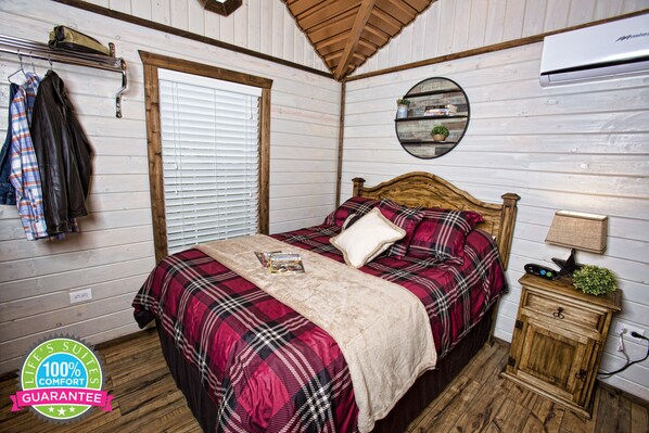 A bedroom with a plaid bedspread, a wooden headboard, a bedside table with a lamp, and a wall-mounted shelf with plants.