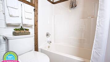 A bathroom with a white toilet, a bathtub, and a shower curtain. There are towels hanging on a rack and a small plant on the toilet.