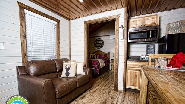 A cozy living room with a brown leather sofa, wooden flooring, a kitchen area with a microwave and refrigerator, and a doorway leading to a bedroom.