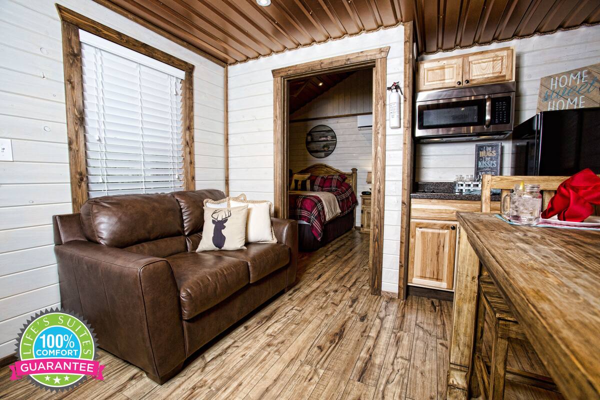 A cozy living room with a brown leather sofa, wooden flooring, a kitchen area with a microwave and refrigerator, and a doorway leading to a bedroom.
