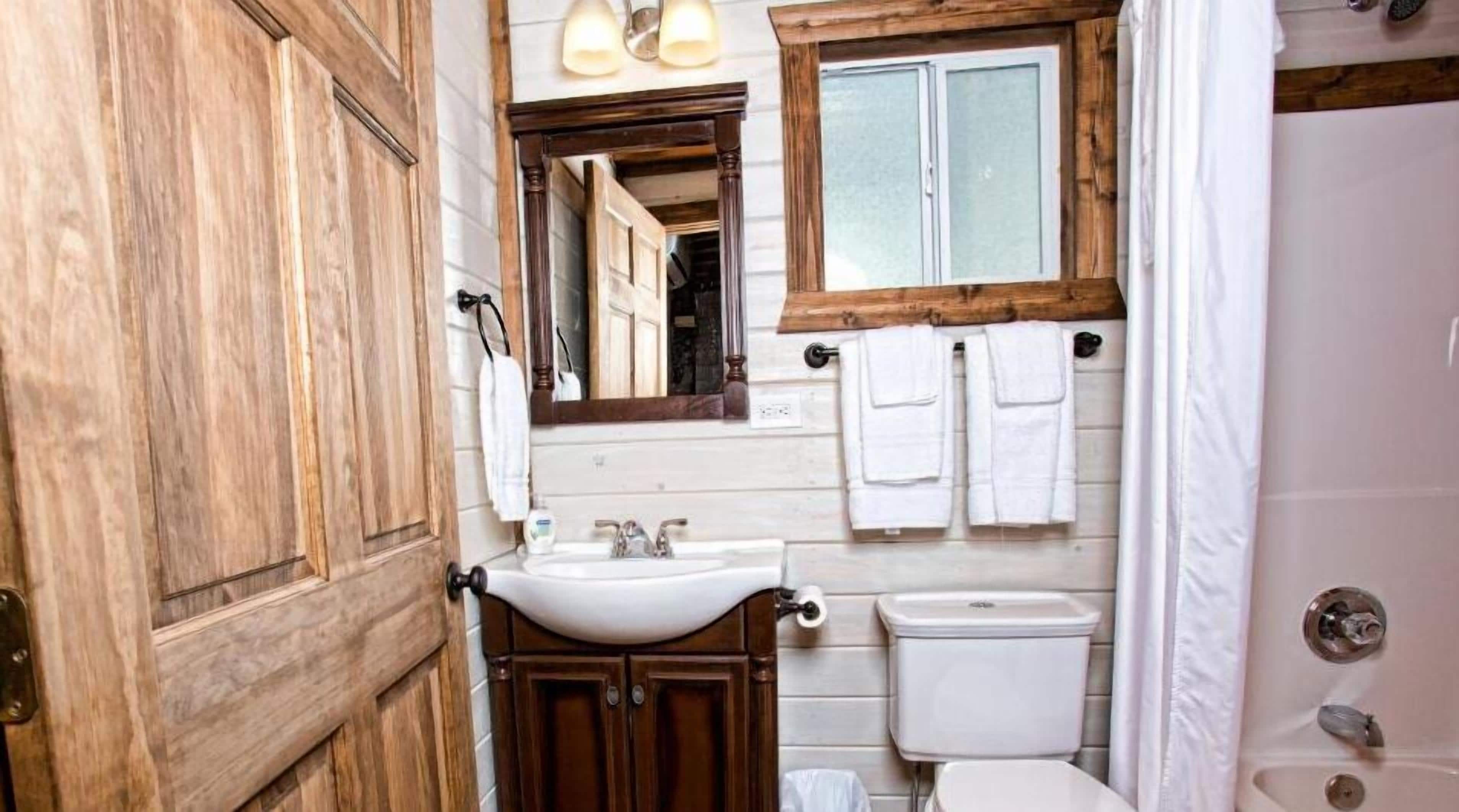 Une salle de bain avec une porte en bois, un lavabo, un miroir, une fenêtre avec un cadre en bois et des toilettes.