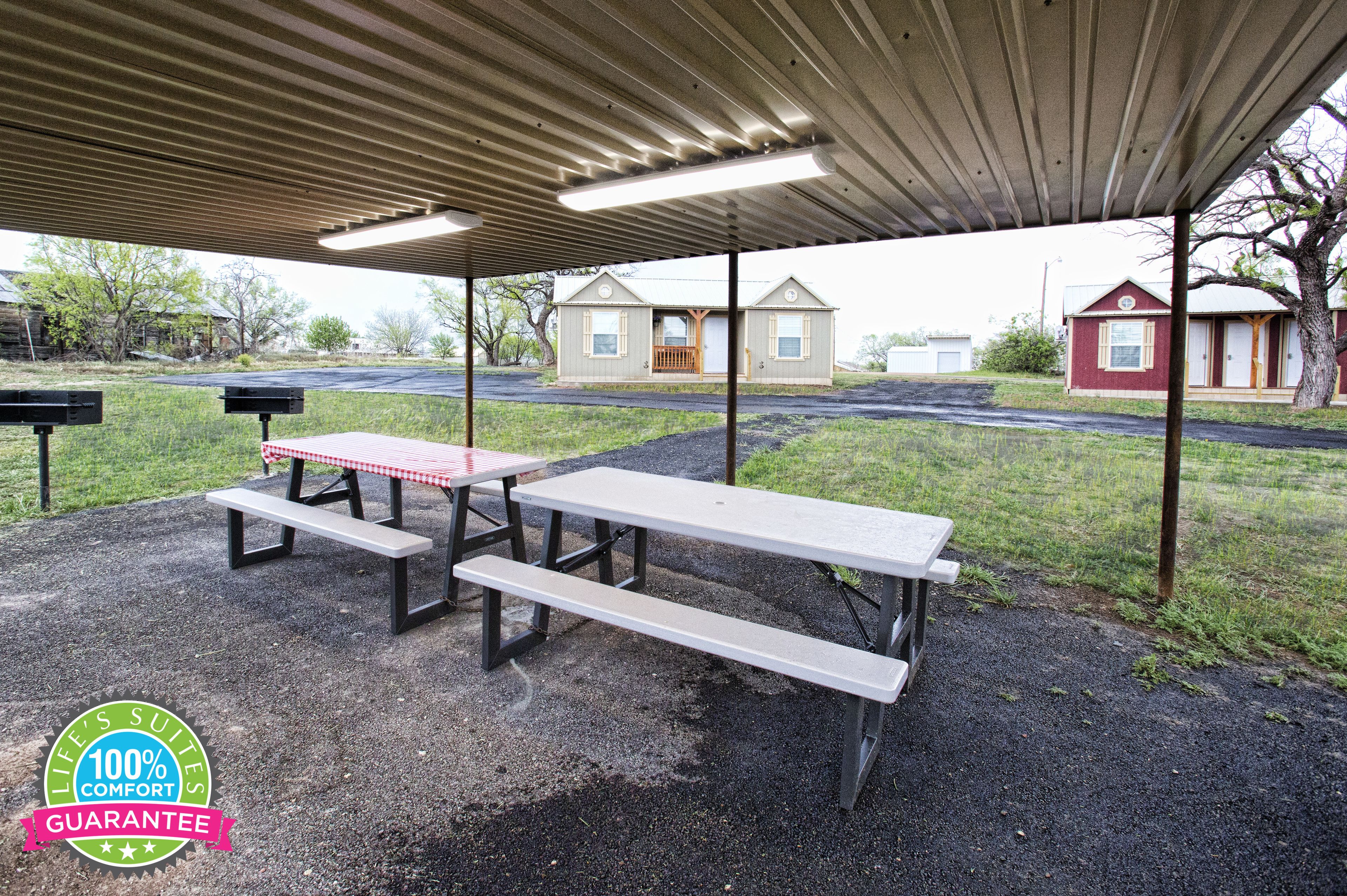 A covered picnic area with benches and picnic tables, a metal roof, and a view of houses in the background.