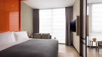 A modern hotel room with a large bed, a TV, a small table with fruit, and a view of the city.