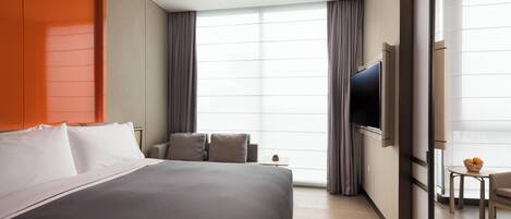 A modern hotel room with a large bed, a TV, a small table with fruit, and a view of the city.
