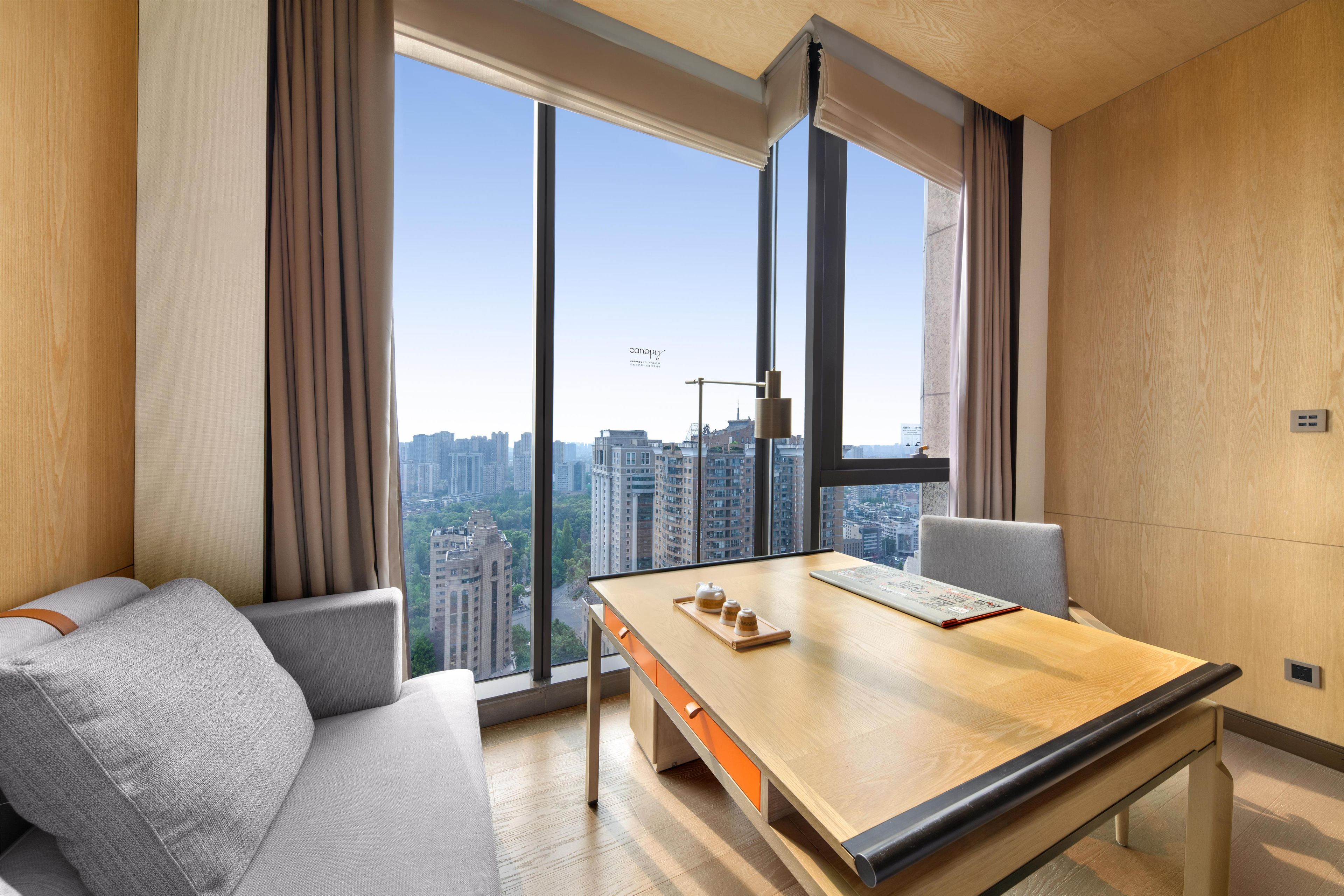 A modern hotel room with a large window, a sofa, a wooden desk, and a city view.