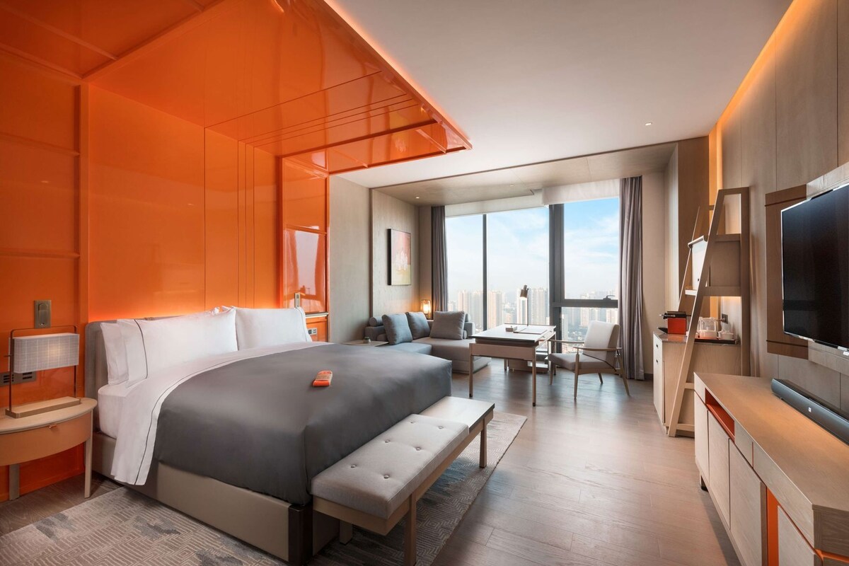 A modern hotel room with a large bed, a desk, a chair, a TV, and a city view.