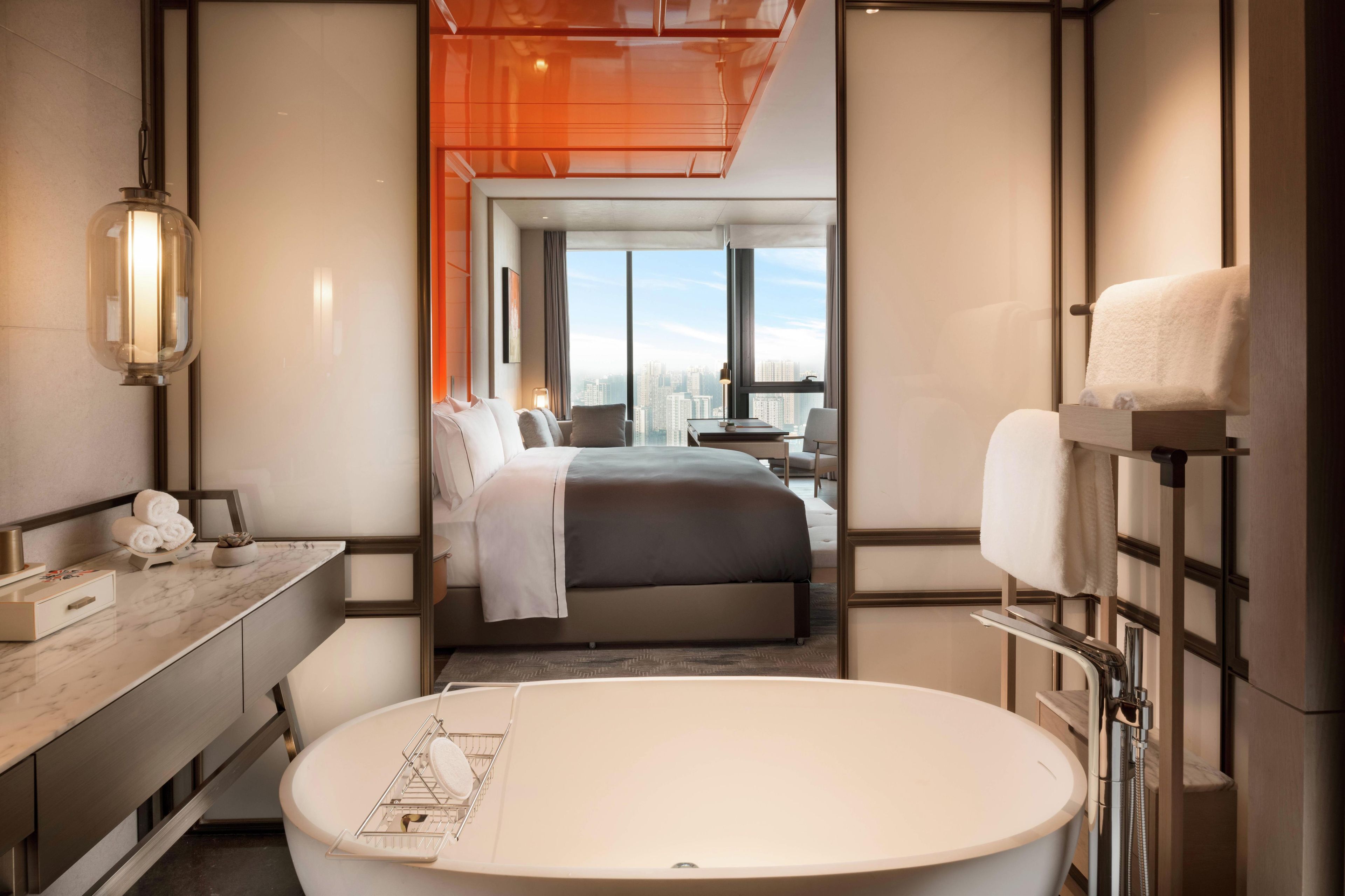 A modern hotel room with a large bed, a round bathtub, a desk with a lamp, and a view of the city.