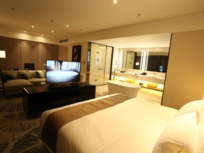 A modern hotel room with a large bed, a flat-screen TV, a sofa, a coffee table, and a bathroom with a bathtub and a large mirror. - Mercure Panjin (Panjin)