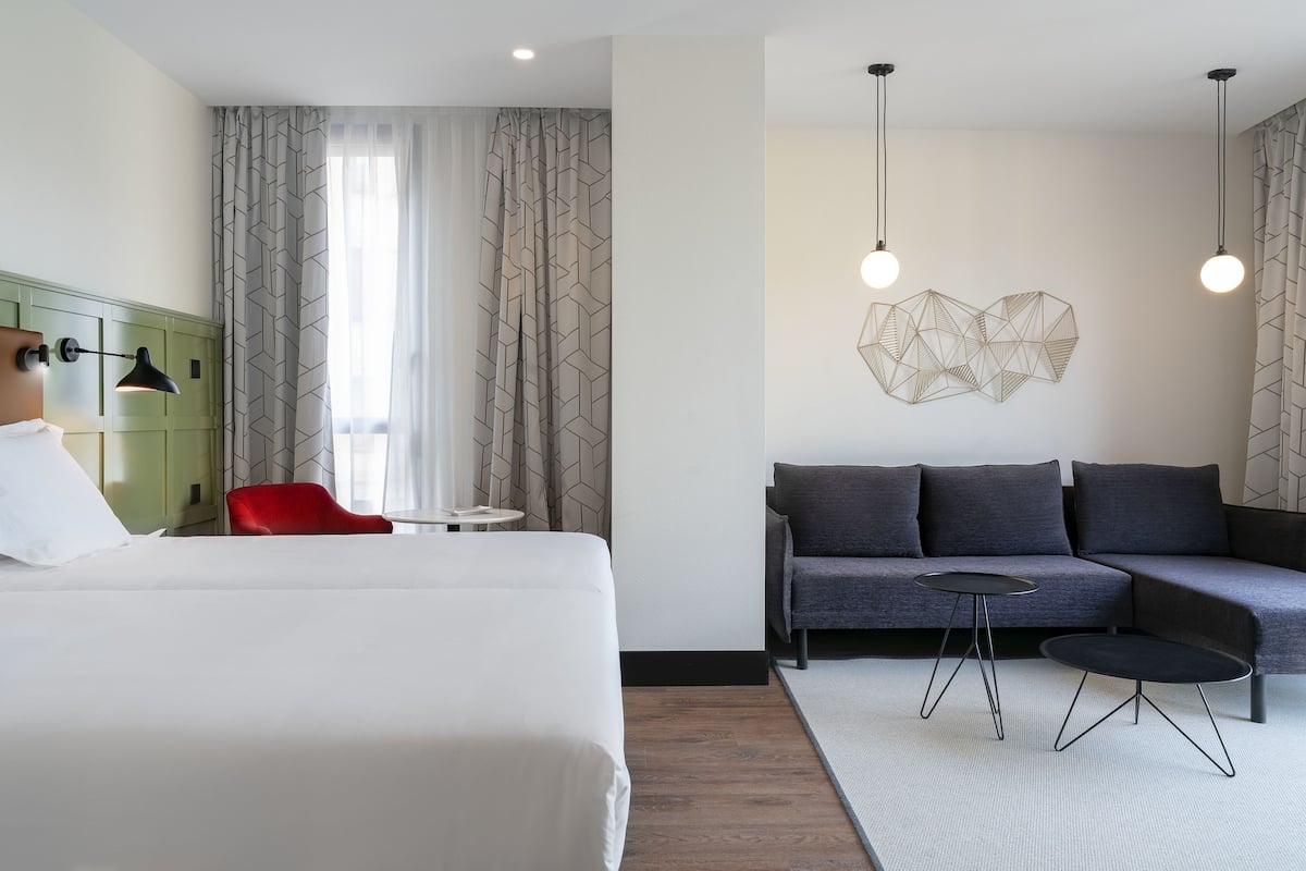 A modern hotel room with a large bed, a grey sofa, two small tables, and a red chair.