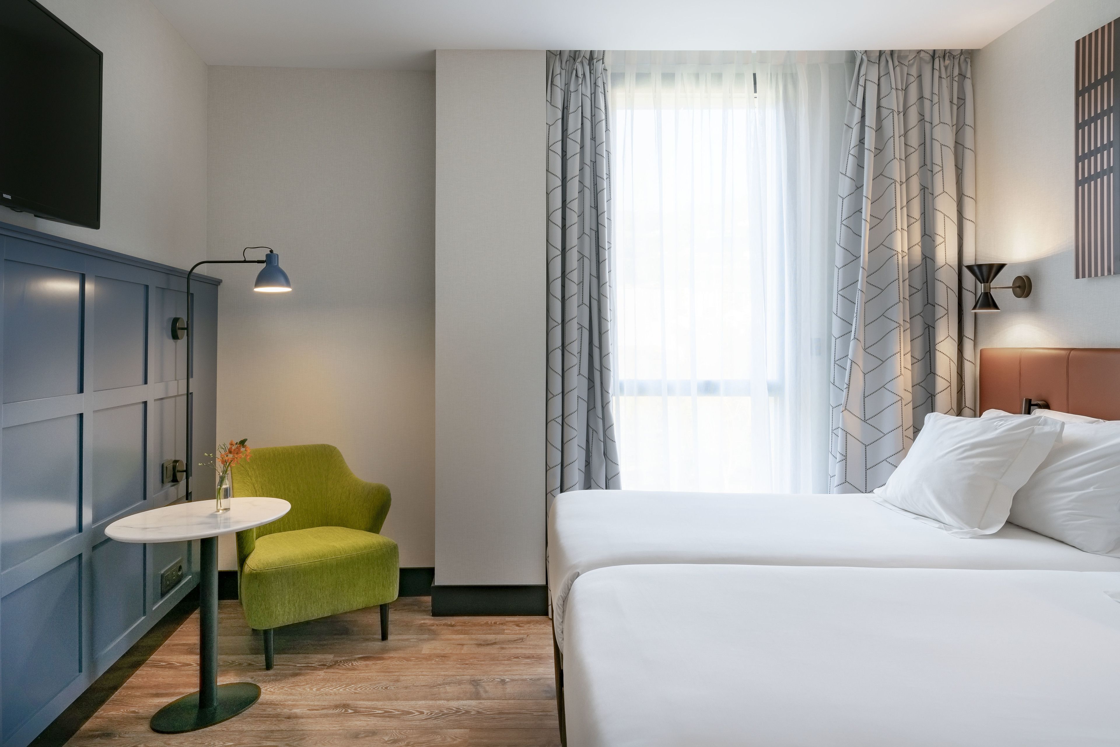 A hotel room with a bed, a green armchair, a small table, and a TV.