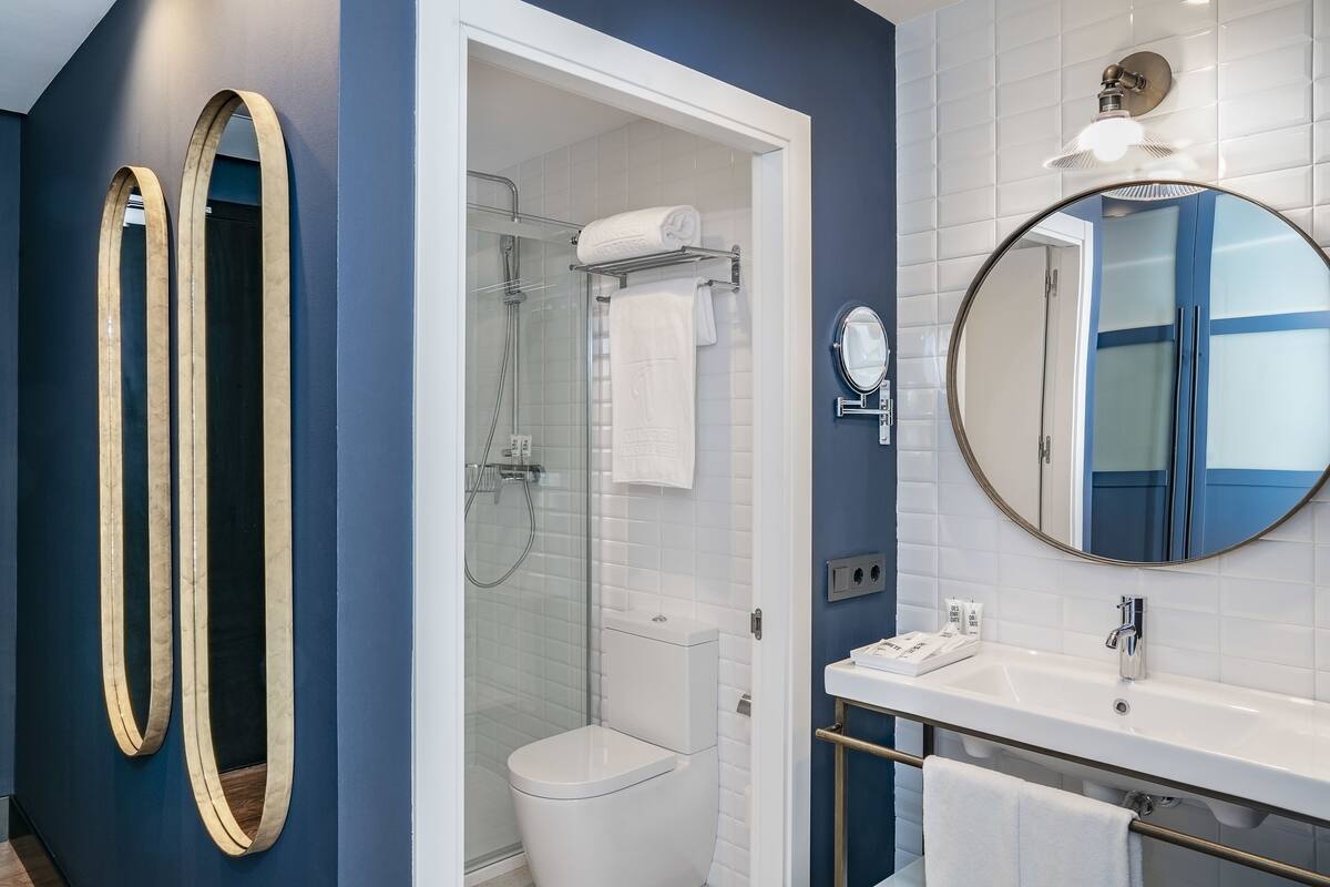 Un baño moderno con ducha de vidrio, inodoro blanco, lavabo con espejo y dos espejos ovalados montados en la pared.