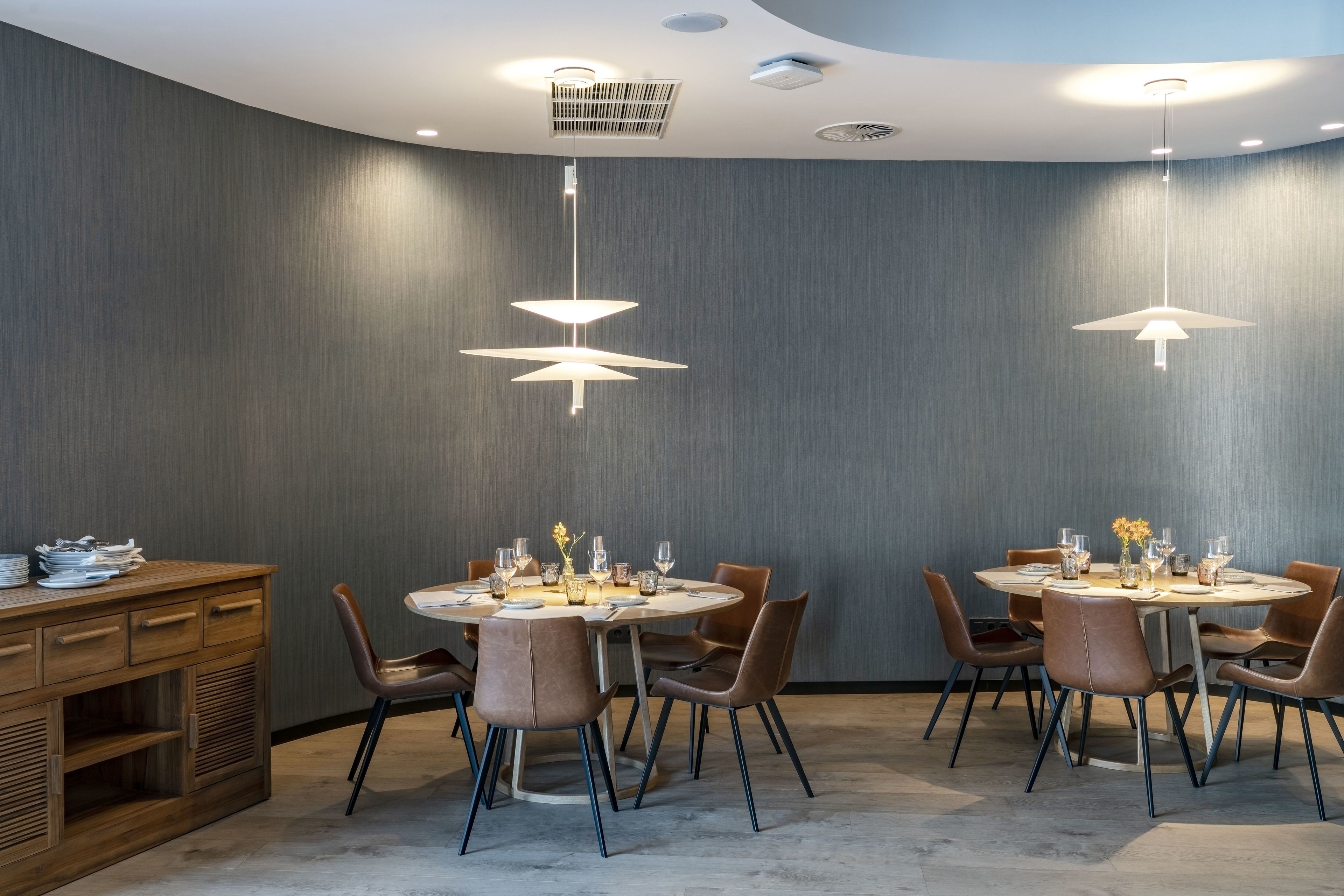 Un comedor moderno con mesas redondas, sillas marrones e iluminación colgante.