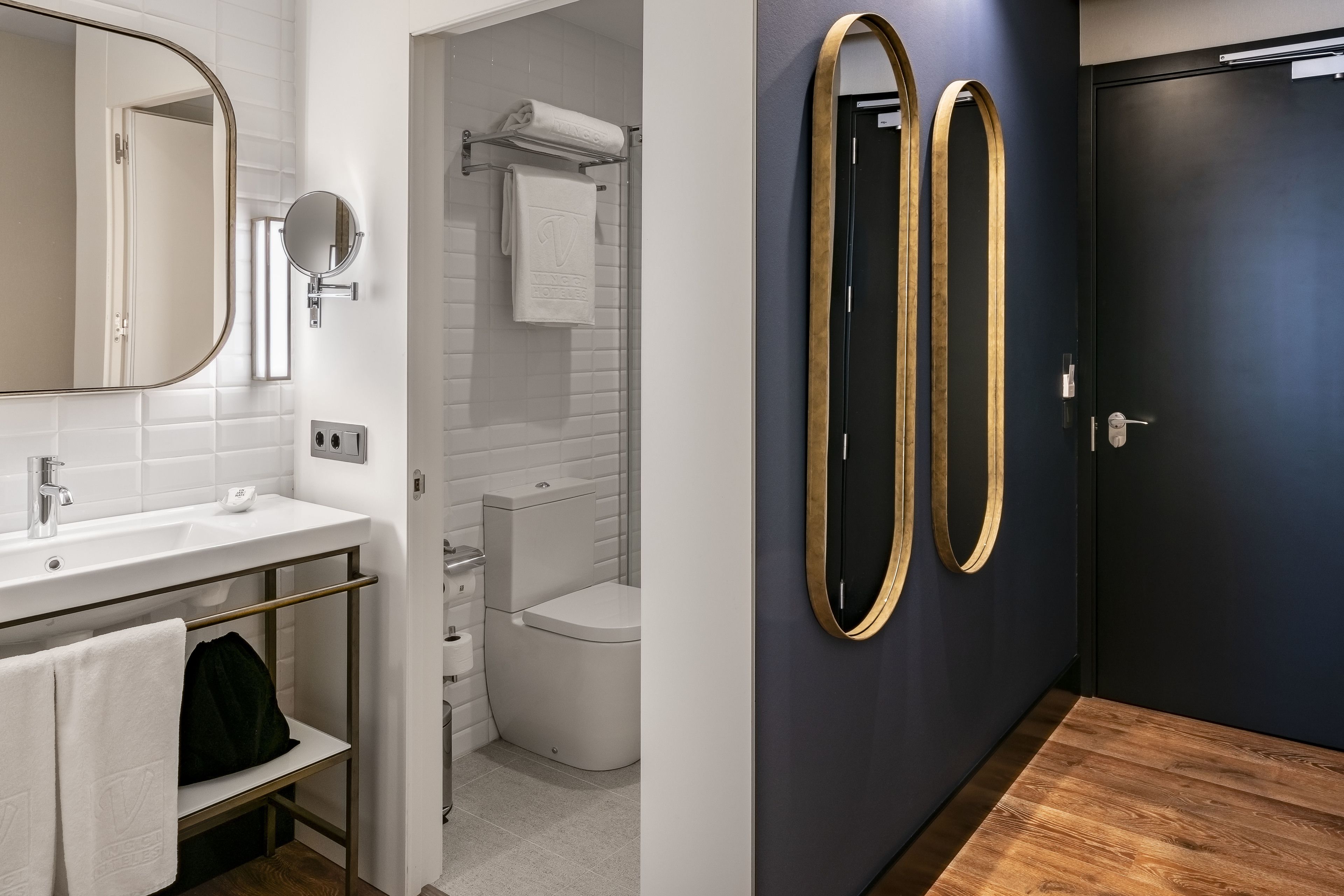 A modern bathroom with a white sink, mirror, and towel rack. There is a dark door and a wooden floor.