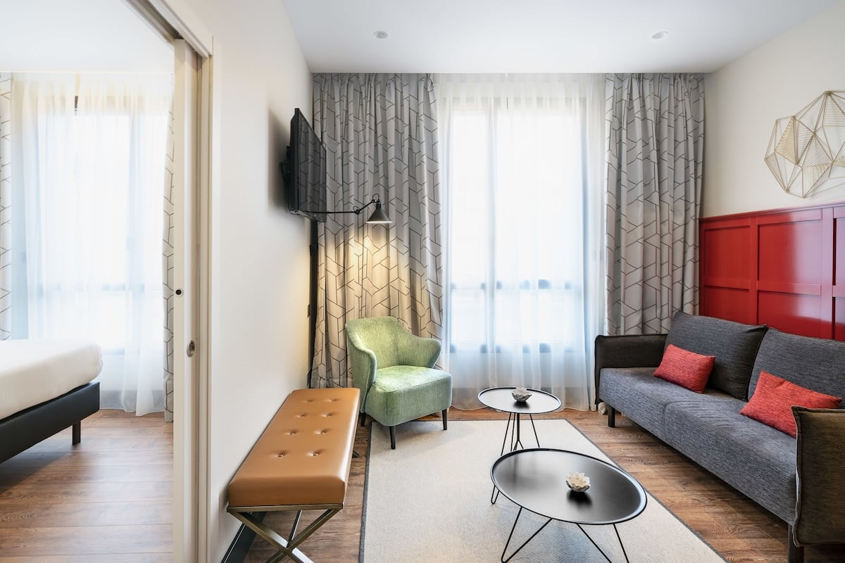 Una habitación de hotel moderna con una cama, un sillón verde, un sofá gris con cojines rojos, una pequeña mesa redonda y un televisor montado en la pared.