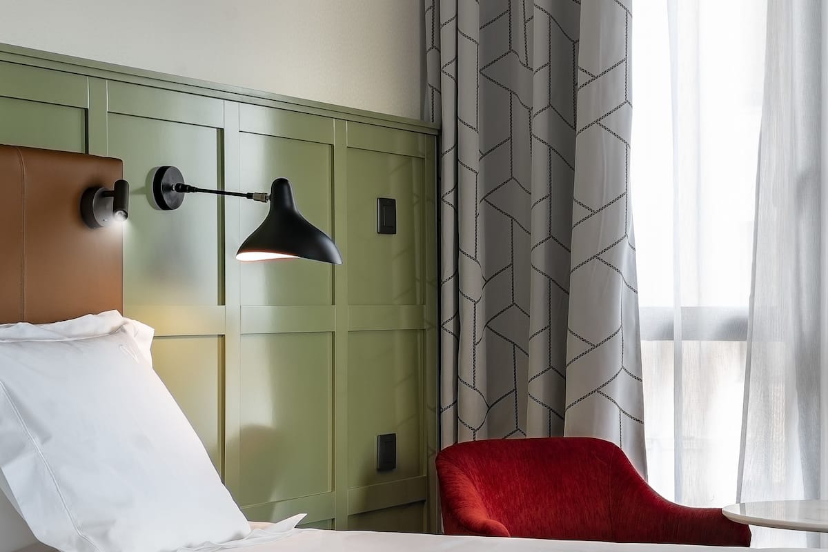 A hotel room with a bed, a red chair, a wall-mounted lamp, and a window with sheer curtains.